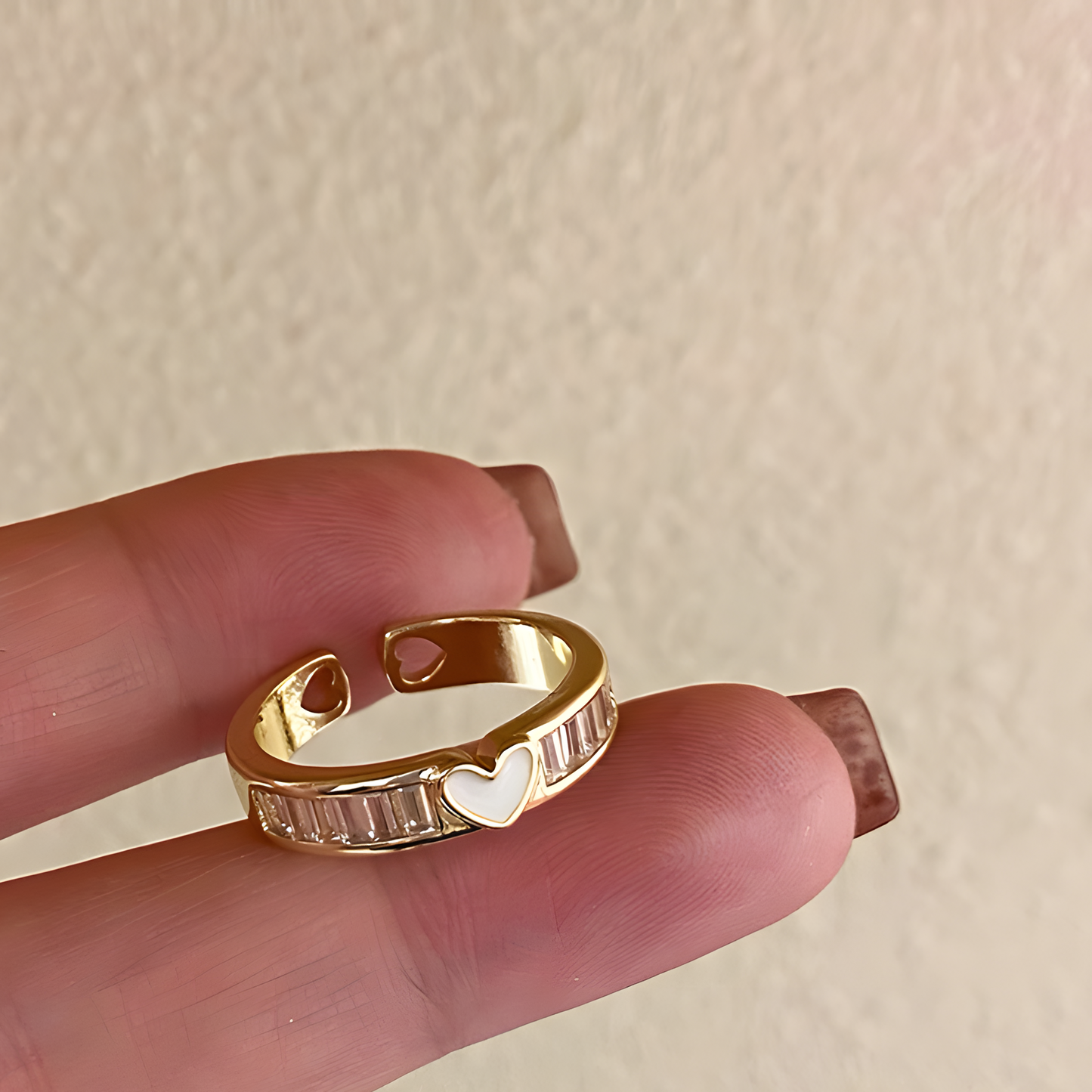 Gold ring with heart detail on a finger against a beige background